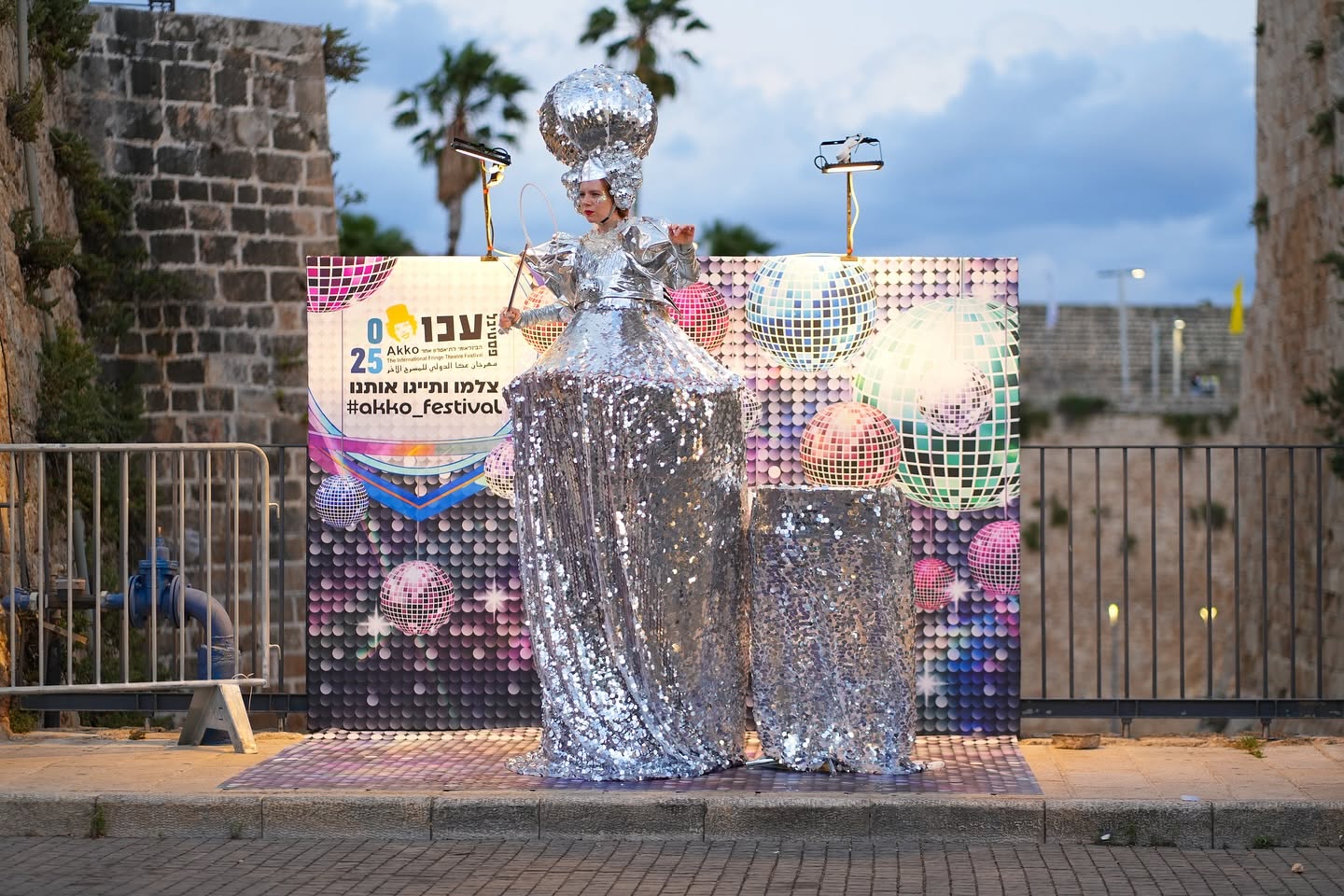 אמנית בתלבושת כסופה ניצבת ברחוב ליד כרזת הפסטיבל.